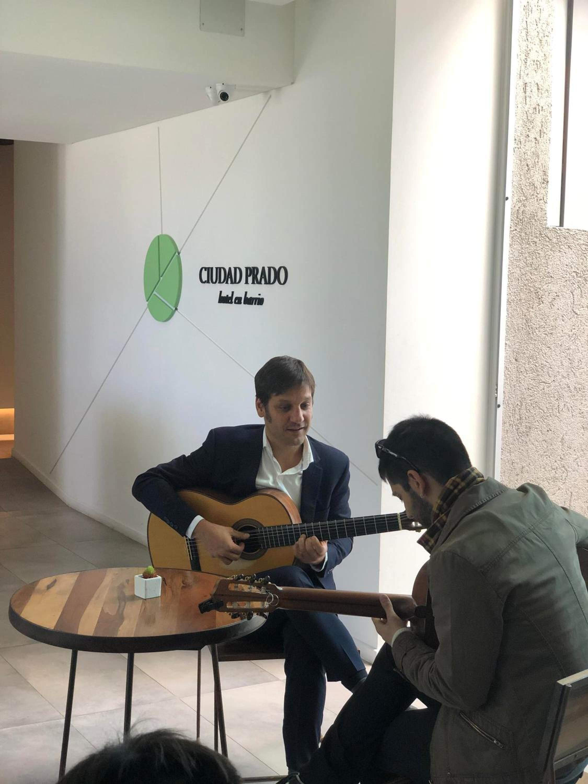 Un hombre tocando la guitarra en una habitación con un reloj de pared y un letrero que dice "CIUDAD PRADO Let the music begin".