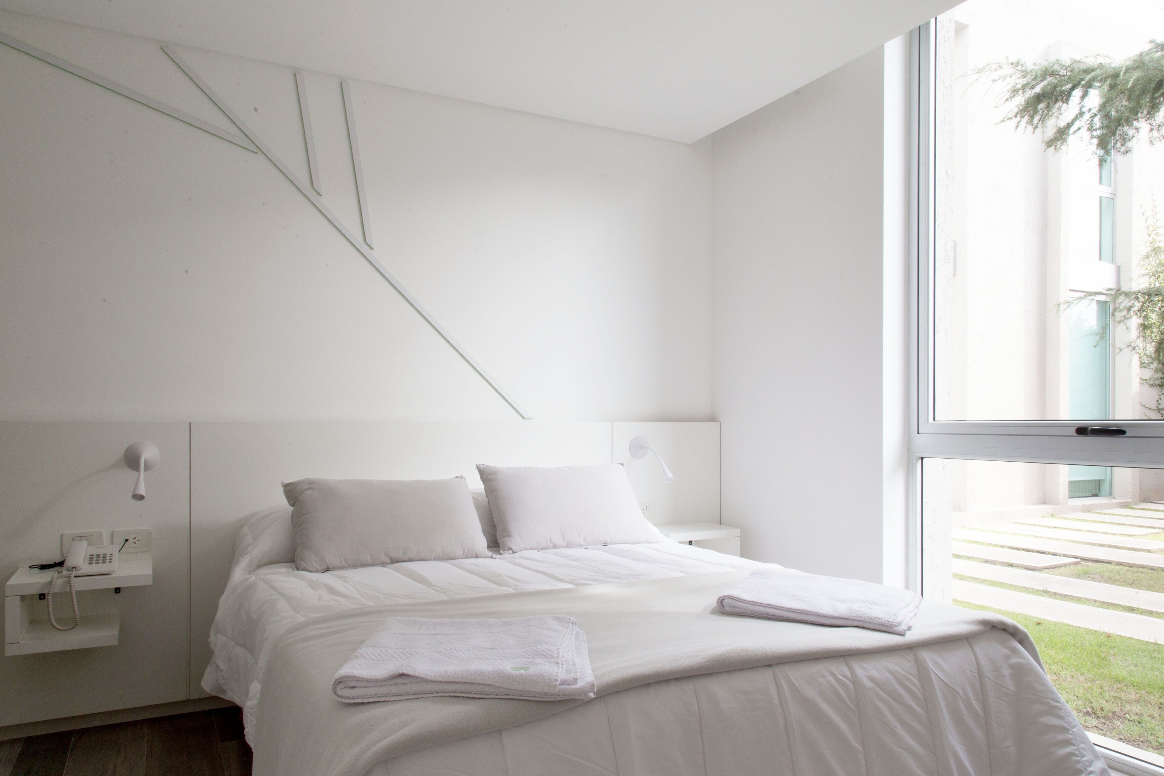 A modern bedroom with a bed, white linens, a bedside table with a lamp, and a window with a view of a building and greenery.