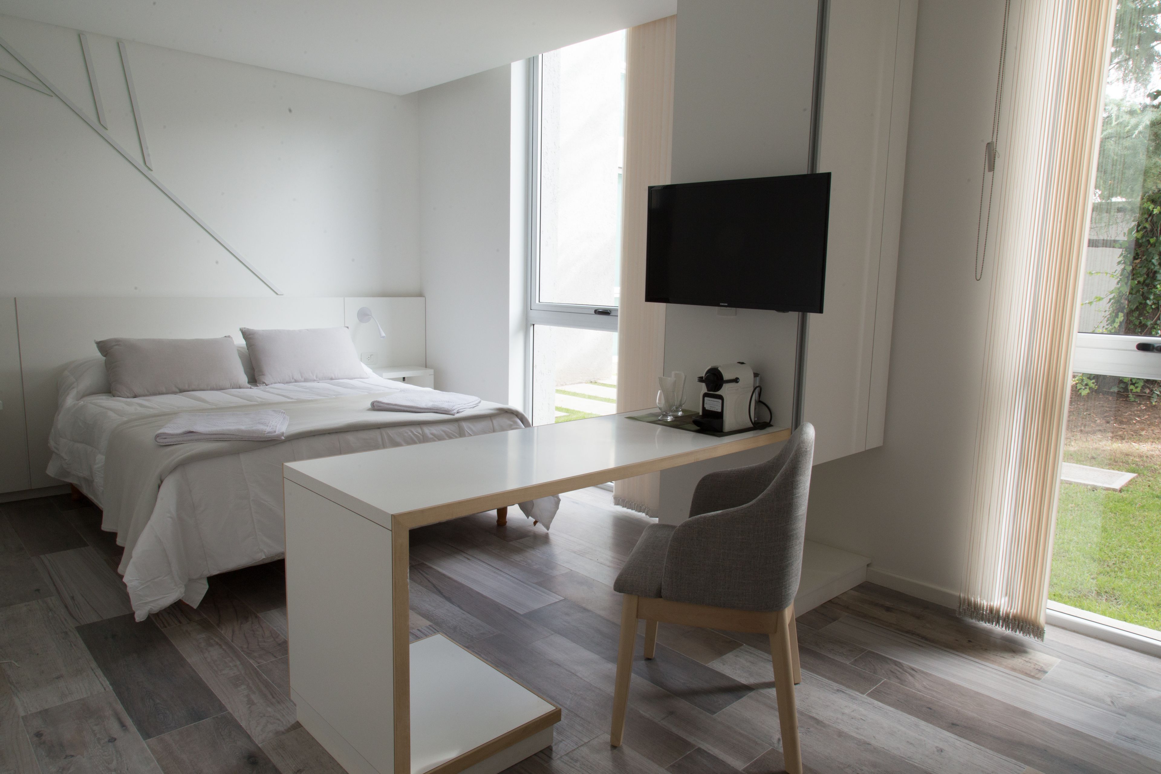 A modern bedroom with a bed, a desk, and a chair. There is a TV mounted on the wall and a window with curtains.