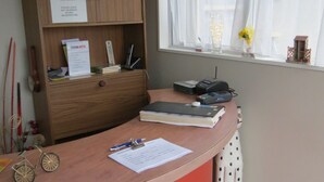 Una taquilla de recepció amb un armari de fusta, un cartell de prohibició de fumar, una finestra amb cortines translúcides i una fotografia encadrada a la paret.