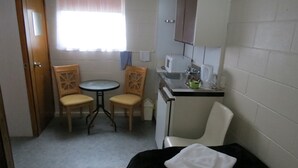 Habitació petita i senzilla amb llit, taula i dues cadires, vàter i finestra amb cortines.