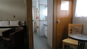 Habitació senzilla amb llit, una cadira de fusta, taula petita i zona de cuina equipada.