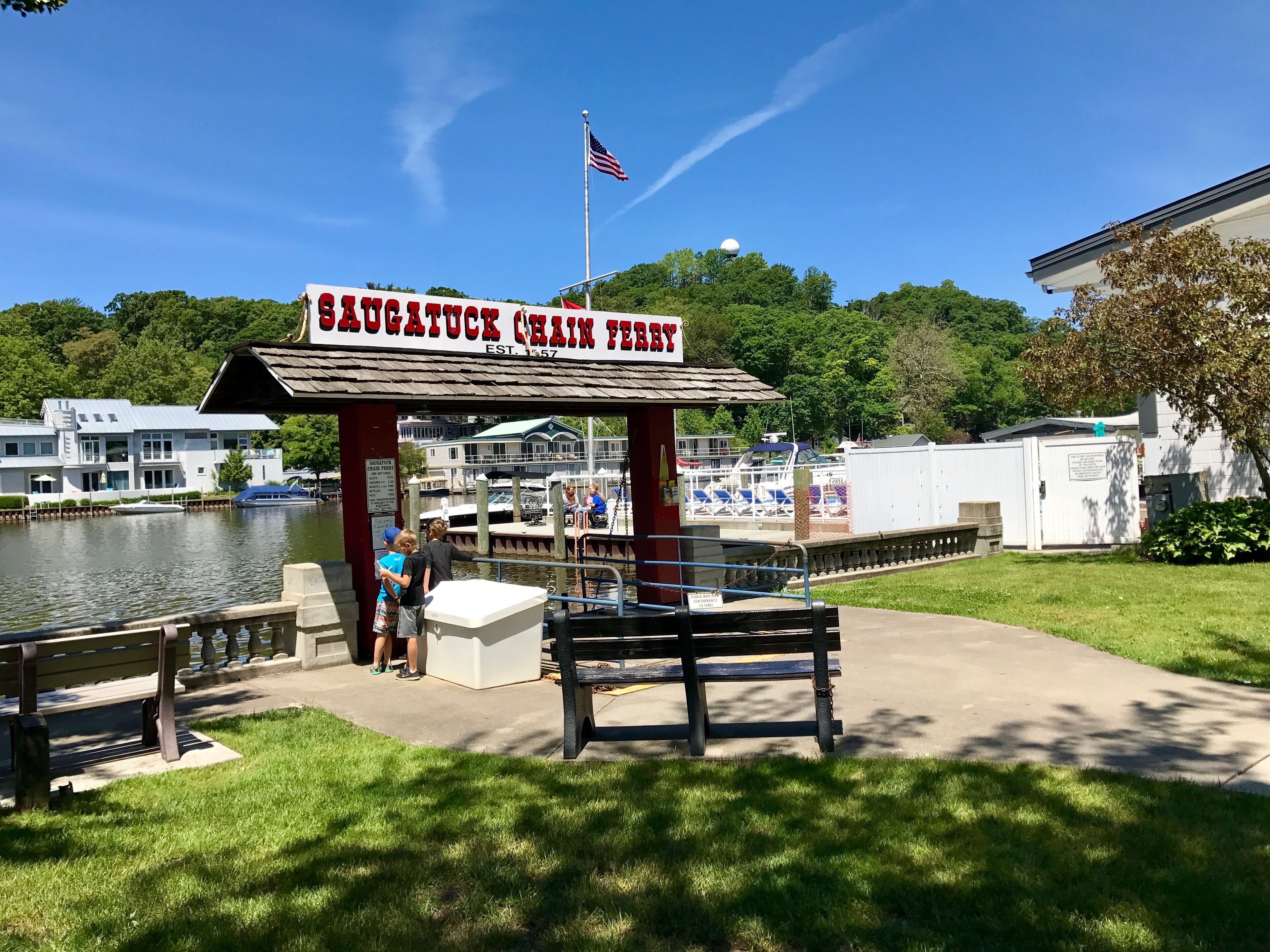 Een restaurant aan het water genaamd 'Saugatuck Chain Ferry', met een steiger, een vlag en een uitzicht op boten en gebouwen op de achtergrond.