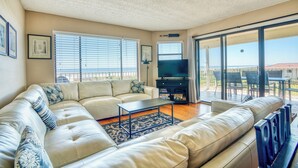 En rummelig stue med stor sofa, sofabord og fjernsyn. Store vinduer med skodder, en balkon med udendørs møbler og et loft med et struktureret mønster.