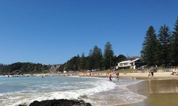 A beach with people swimming and walking along the shore, surrounded by trees and buildings. - SALT by the Sea - Unit 1 (Port Macquarie)