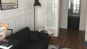 Un salon avec un canapé noir, une petite table avec une lampe, un tapis à motifs géométriques et une citation encadrée accrochée au mur.