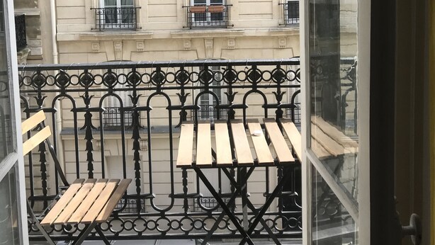 Un balcon doté d’une table et de chaises en bois, d’une balustrade en fer forgé et offrant une vue sur un immeuble avec de nombreuses fenêtres et des balcons.