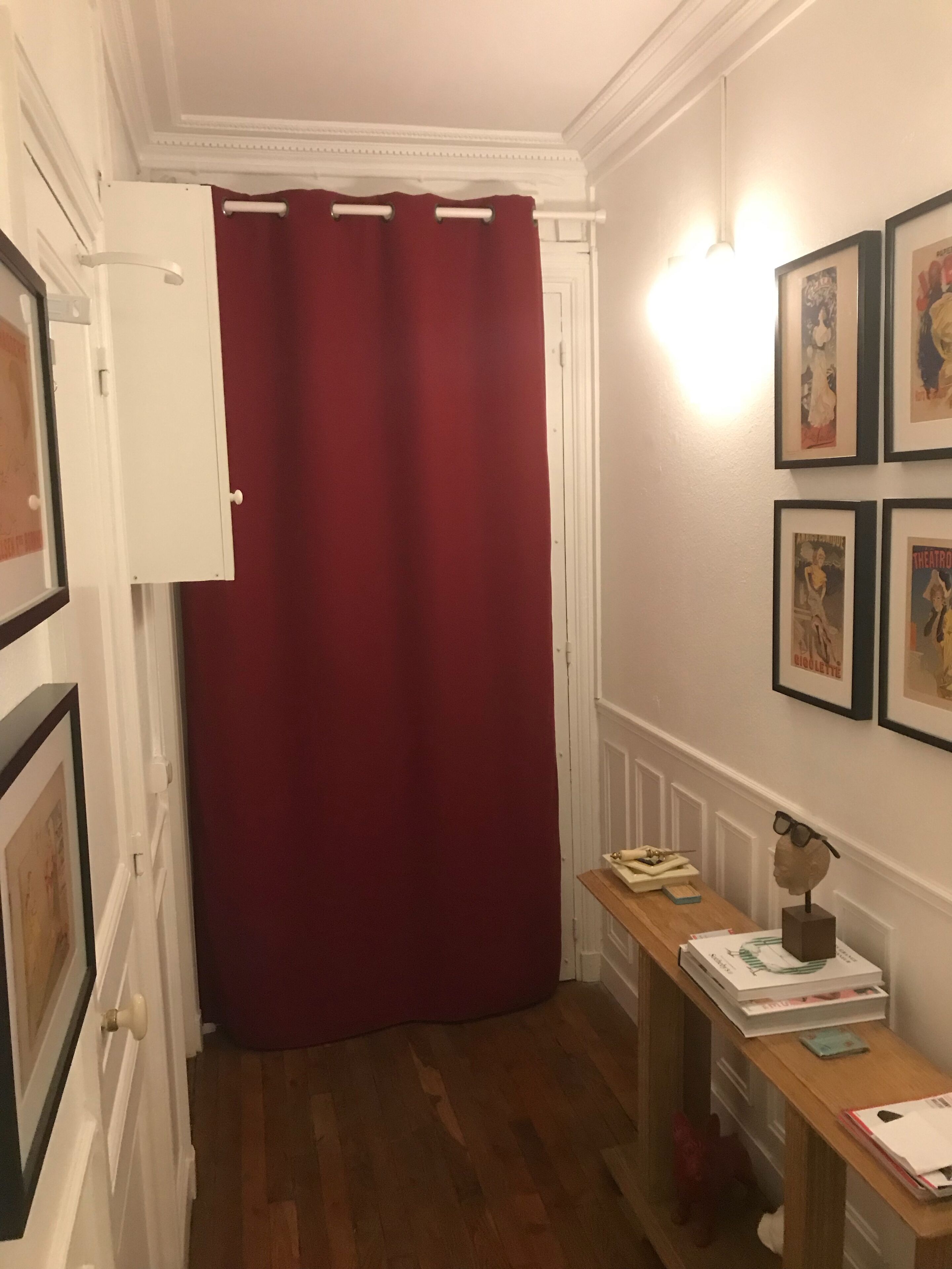 Ein Flur mit rotem Vorhang, gerahmten Bildern an der Wand, einem Holztisch mit Büchern und einem kleinen dekorativen Gegenstand.