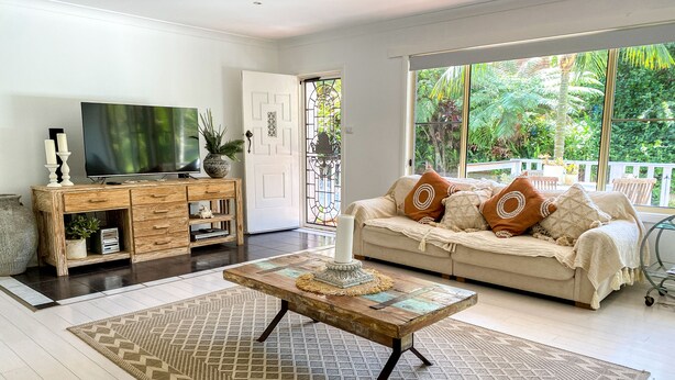 Una sala de estar con un mueble de madera para el entretenimiento, un sofá color beige con cojines estampados, una mesa de centro y un ventanal con vistas a la vegetación.