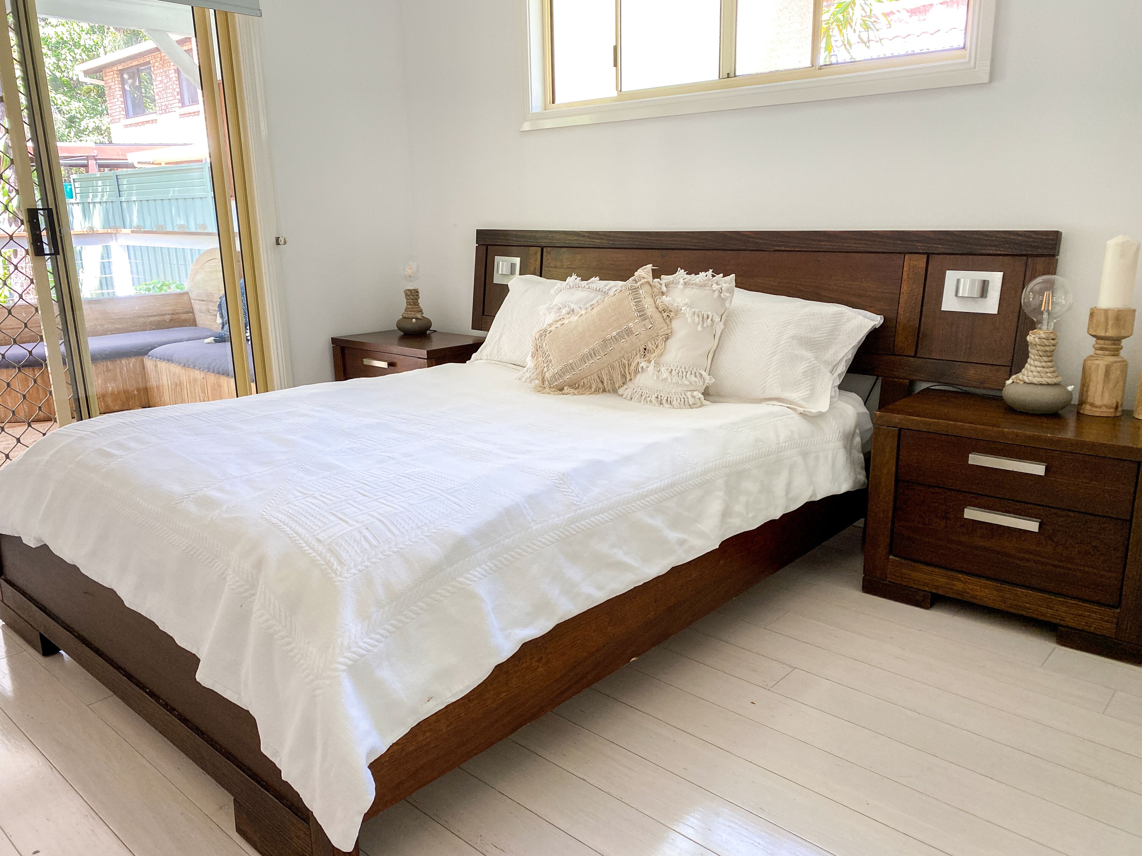 Une chambre à coucher avec un lit en bois, du linge de lit blanc, une table de chevet avec une lampe et une fenêtre donnant sur l’extérieur.