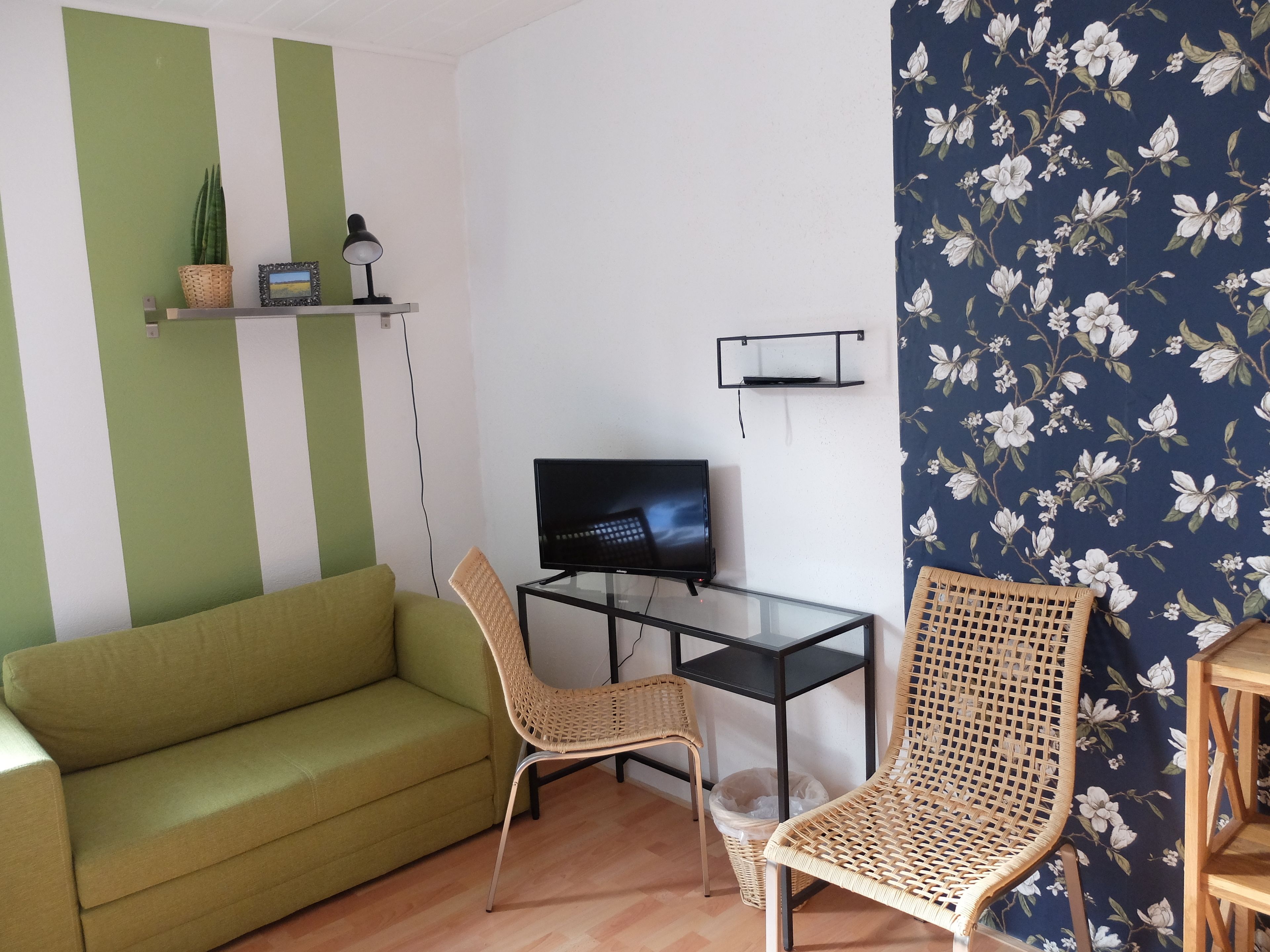 A living room with a green sofa, two wicker chairs, a glass TV stand, and a wall with a floral pattern.