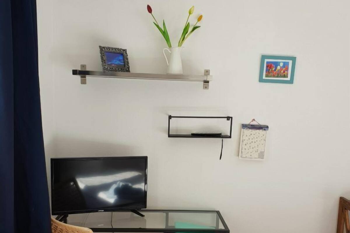 A small, tidy room with a glass desk, a television, a wicker chair, and a small shelf with a vase of flowers.