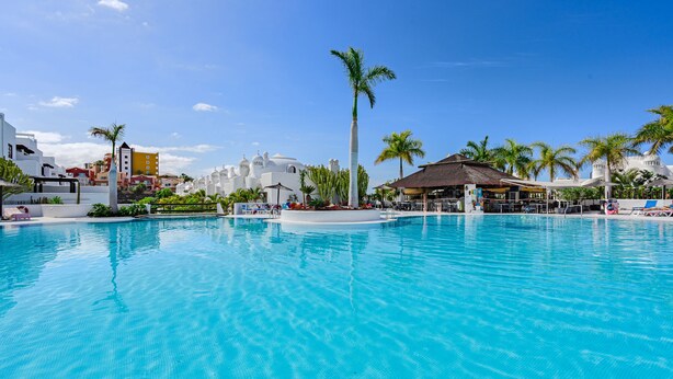 A large swimming pool with clear blue water, surrounded by palm trees and a white building with a thatched roof. - Adeje paradise Duplex with sea view (adeje)