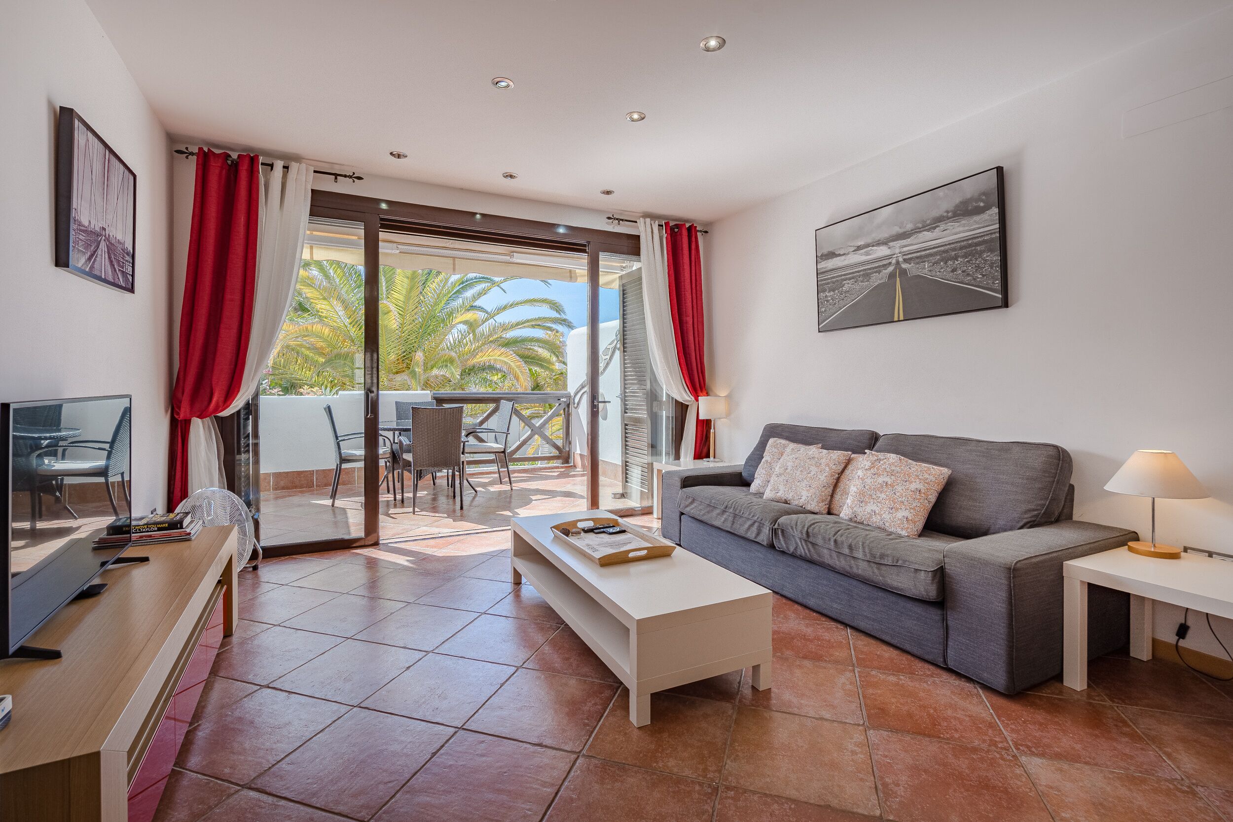 Una sala moderna con un sofá gris, una mesa de centro blanca y un televisor sobre un mueble de madera. Cuenta con un balcón con cortinas rojas y vistas a palmeras.