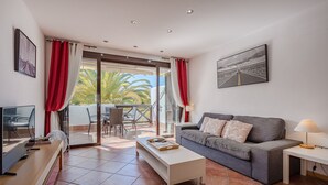 A modern living room with a grey sofa, a white coffee table, and a TV on a wooden stand. There is a balcony with red curtains and a view of palm trees. - Adeje paradise Duplex with sea view (adeje)