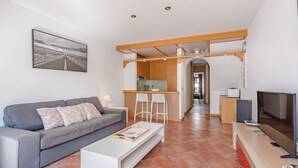 A modern living room with a grey sofa, white coffee table, and a TV on a wooden stand. A kitchen area is visible in the background with a bar stool and a refrigerator. - Adeje paradise Duplex with sea view (adeje)