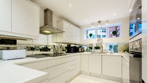 A modern kitchen with white cabinetry, a stainless steel range hood, and a large window providing natural light. - Village Serenity, by Veeve (London)