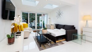 A modern living room with a sofa, coffee table, and TV. There are windows with a view of greenery outside. - Village Serenity, by Veeve (London)