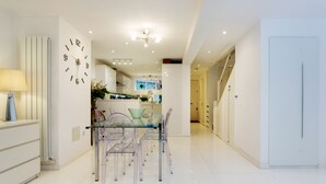 A modern kitchen with white cabinetry, a glass dining table with chairs, and a wall clock. - Village Serenity, by Veeve (London)