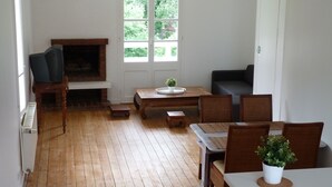 A living room with a fireplace, wooden flooring, a dining table with chairs, and a large window with a view of greenery. - Magnificent apartment (80 m²) with 4 main rooms, 100 meters from the beach (Hermanville-Sur-Mer)