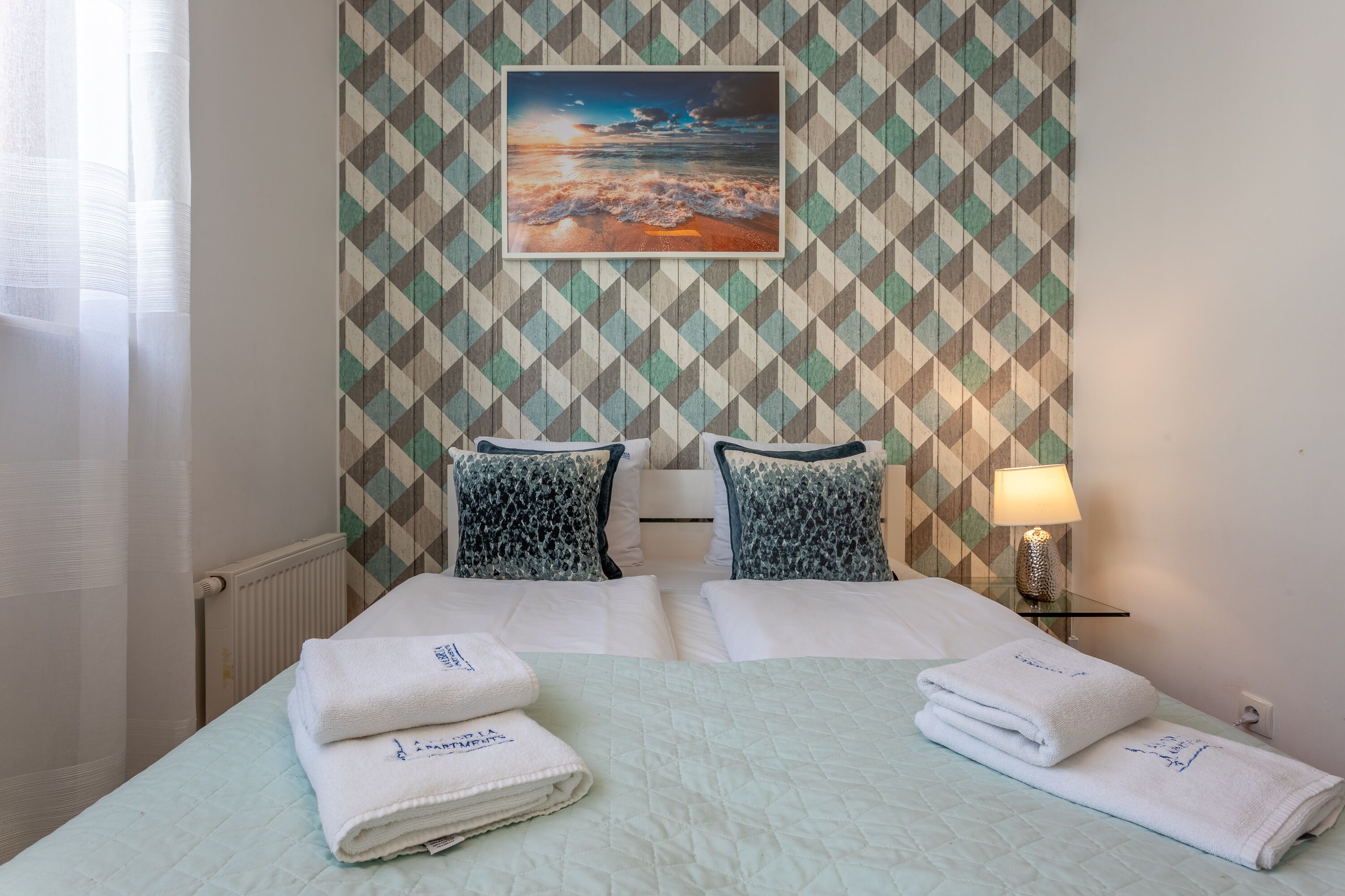 A hotel room with a bed, two pillows, a nightstand with a lamp, and a wall with a geometric pattern.