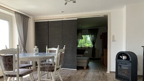 Et spiseområde med et bord og stole, en sort pejs og udsigt til en have gennem et vindue.