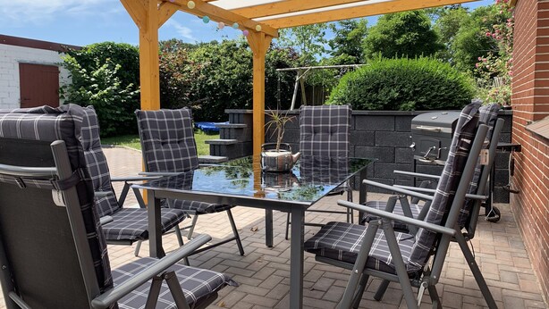 En udendørs terrasse med et glasbord og stole, en pergola med lyskæder, samt en murstensvæg.