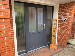 Une porte de couleur sombre avec un panneau en verre dépoli, une enseigne en bois portant une inscription et un décor en forme de cœur sur le mur.