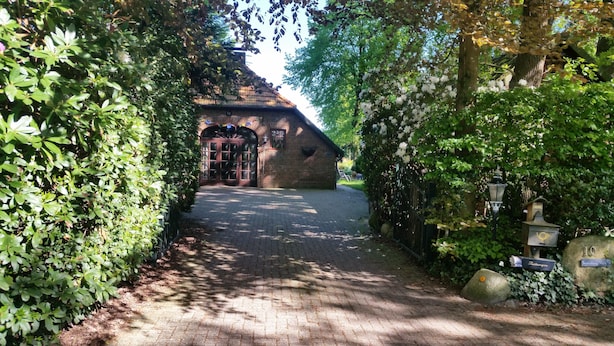 Un bâtiment en briques à l’architecture traditionnelle, doté d’une entrée pavée et entouré d’une végétation luxuriante.