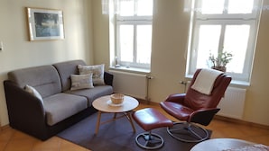 A living room with a grey sofa, a red leather chair, a small round table, and a window with lace curtains.