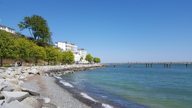 A coastal scene with a rocky shoreline, a pier extending into the water, and buildings with balconies overlooking the sea.