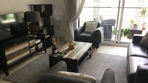 Un salón moderno con un televisor de pantalla plana, una mesa de centro con una botella y vasos, un sofá y una ventana que ofrece vistas al exterior.