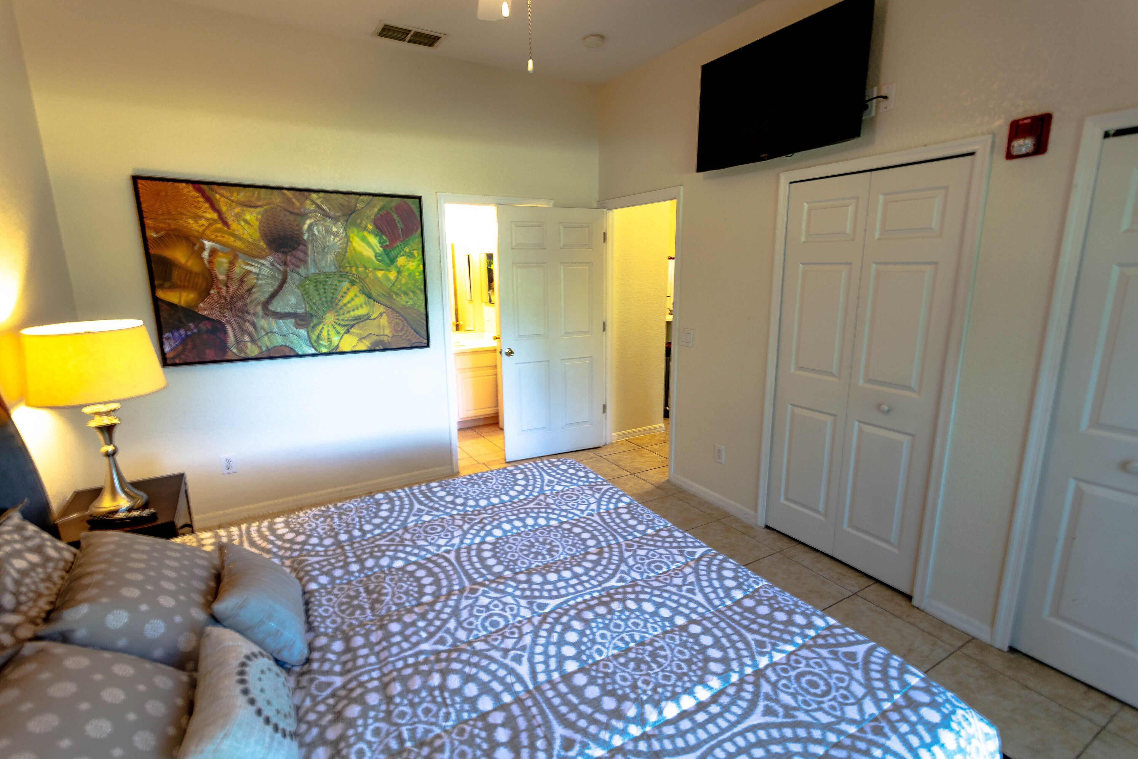 Une chambre à coucher avec une couvre-lit à motifs, un tableau accroché au mur et un téléviseur fixé au-dessus d’une porte.