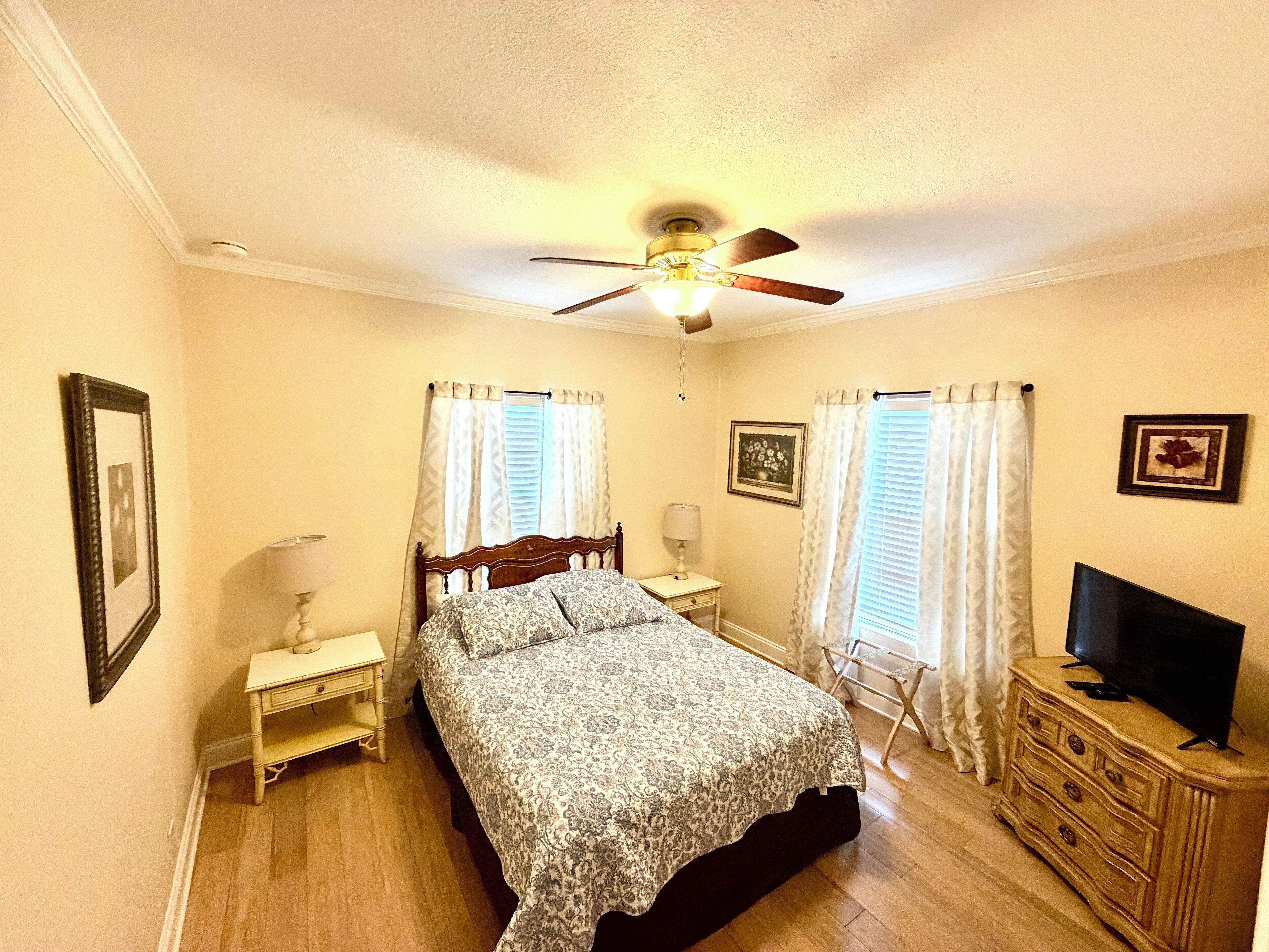 A bedroom with a bed, a ceiling fan, a nightstand, a dresser, a television, and two windows with curtains.