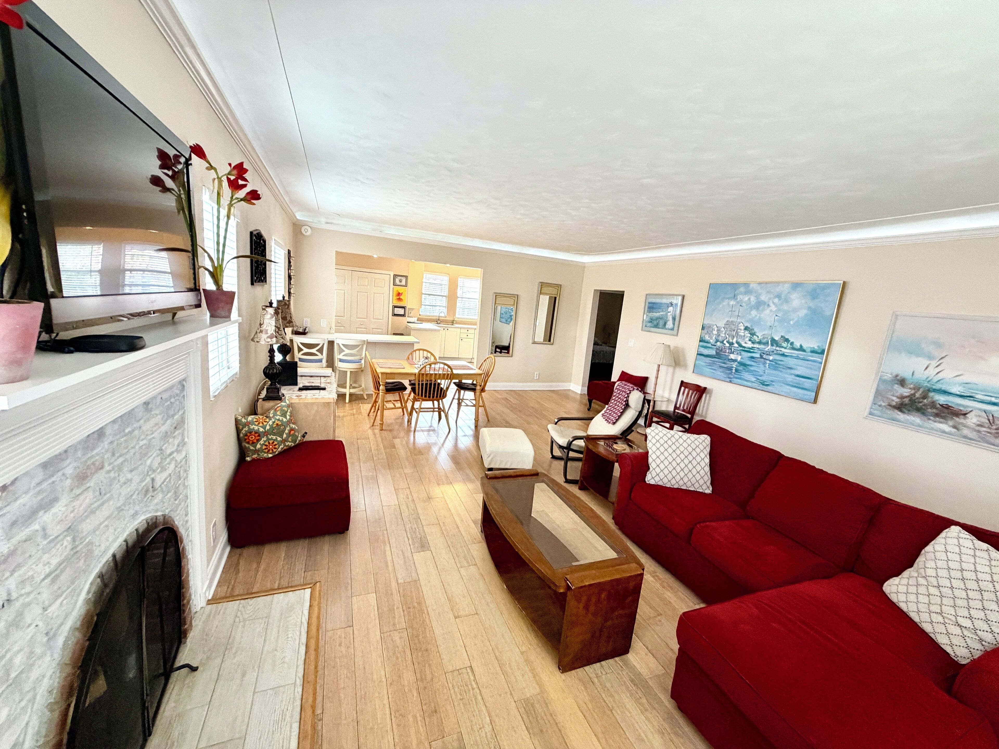 A spacious living room with a red sofa, a wooden coffee table, and a fireplace. There is a dining area with a table and chairs, and a kitchen in the background.
