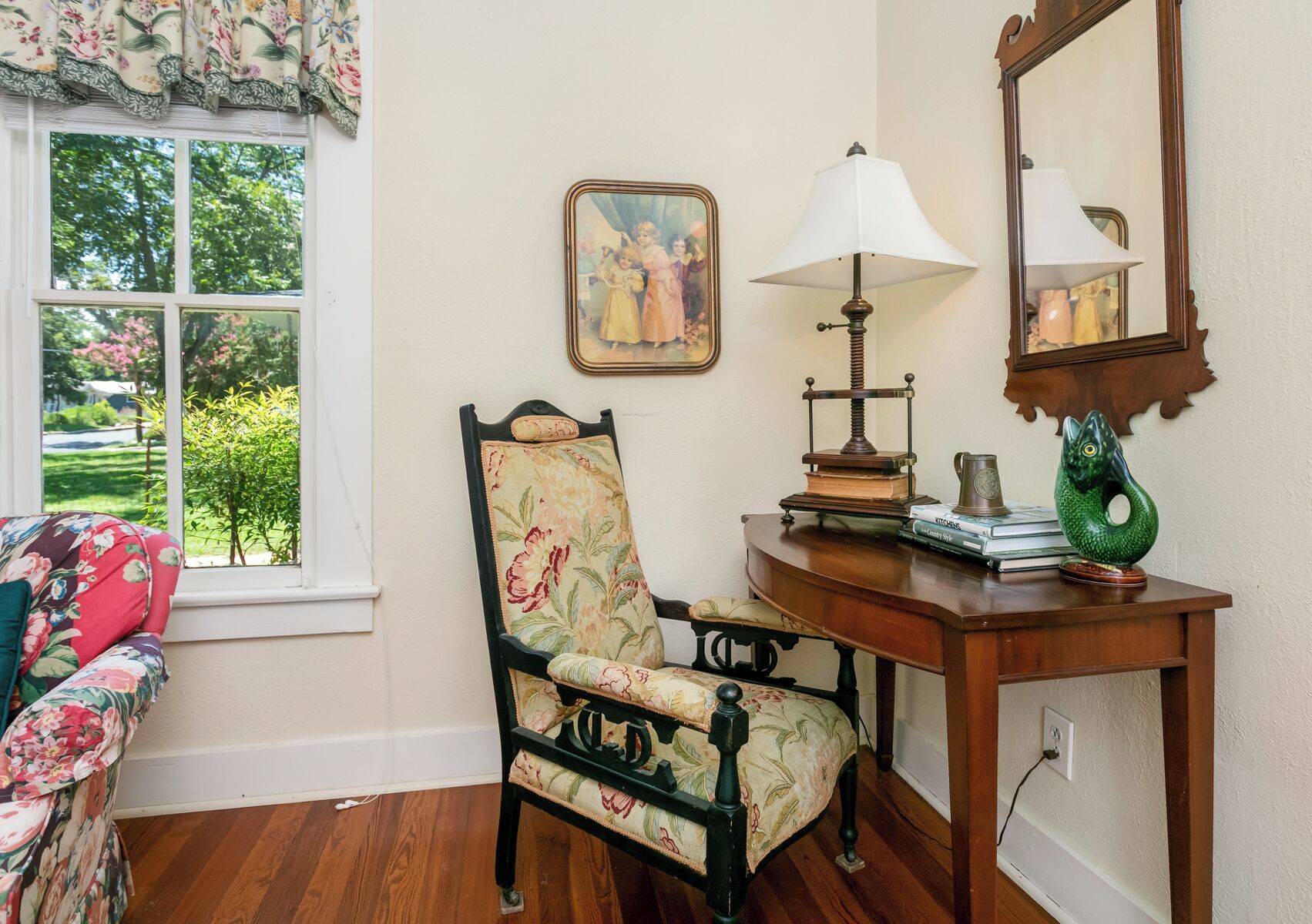 A room with a floral-patterned armchair, a wooden side table with a lamp, a framed painting, and a window with a view of greenery.