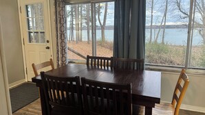 Un comedor con una mesa y sillas de madera, una ventana con cortinas, y vistas a árboles y agua.
