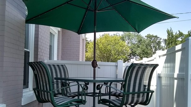 A patio with a table and four chairs, an umbrella, and a brick house in the background. - Truly Unique! 1938 Historic Beach House (Carolina Beach)
