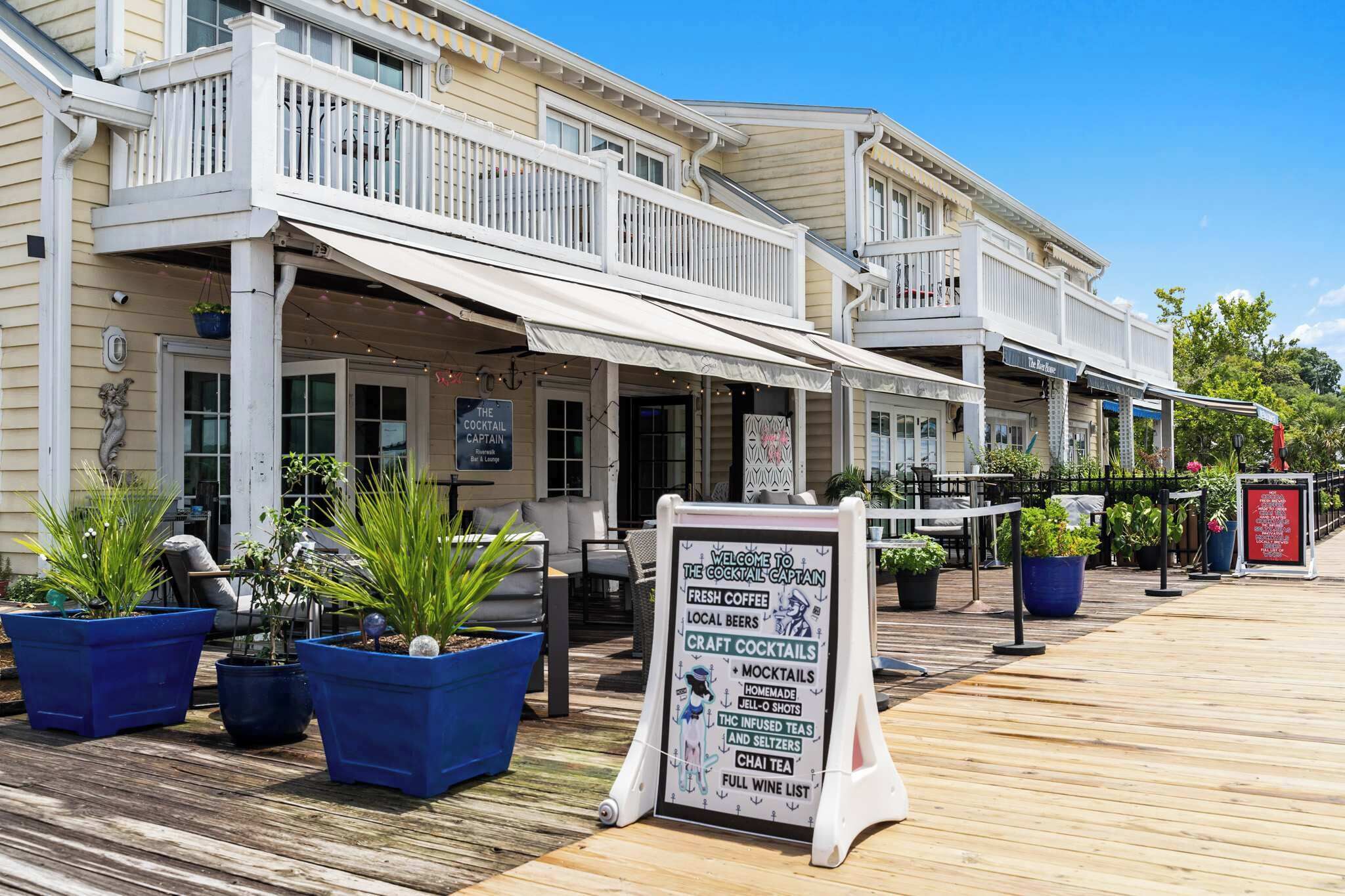 A waterfront bar with outdoor seating, a signboard listing offerings like fresh coffee and craft cocktails, and a wooden deck.