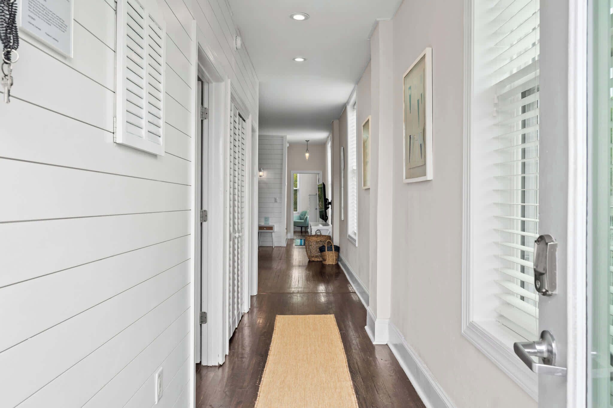 A hallway with white walls, a wooden floor, and a beige rug. There are doors on both sides, and a window on the right.