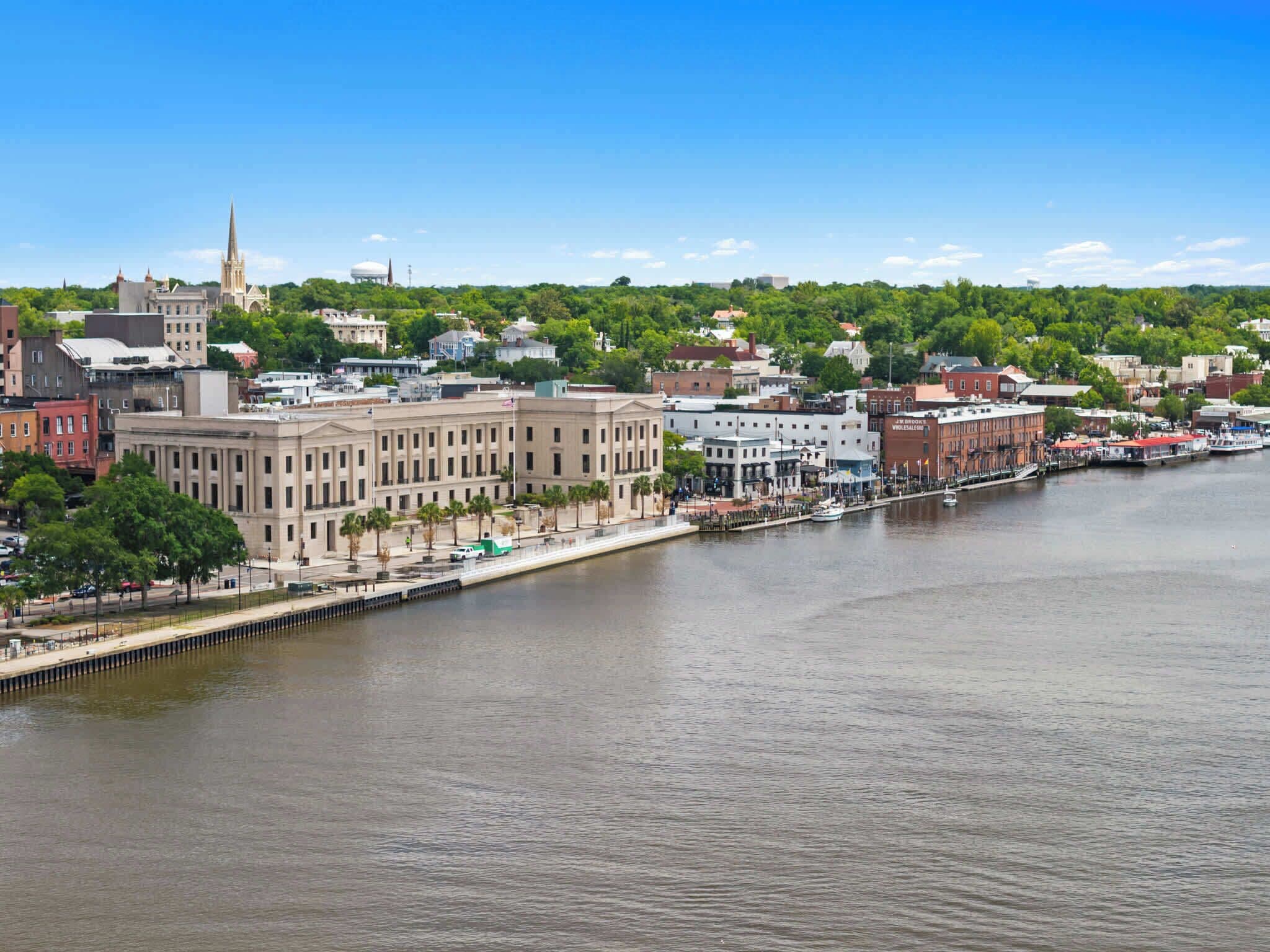 A riverside cityscape with historic buildings, a prominent church spire, and a waterfront promenade.