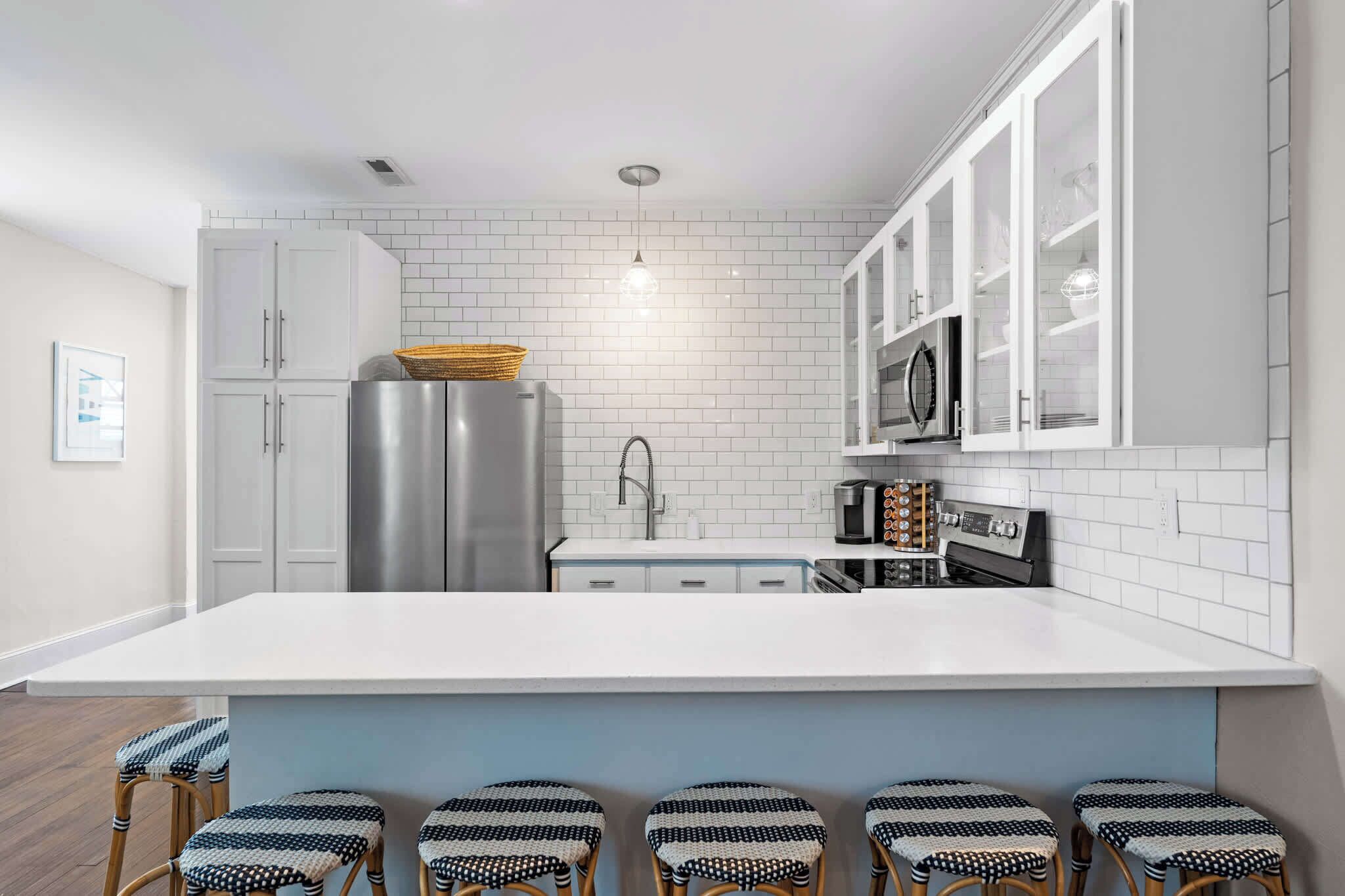 A modern kitchen with white cabinetry, a stainless steel refrigerator, a microwave, and a coffee machine. There are five bar stools with a woven seat.