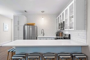 Een moderne keuken met witte kastjes, een roestvrijstalen koelkast, een magnetron en een koffieapparaat. Er zijn vijf barkrukken met een geweven zitvlak.