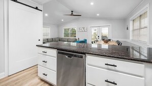A modern kitchen with white cabinetry, a black granite countertop, and a dishwasher. There is a ceiling fan and a window with blinds. - 1min Walk To The Beach, Private Heated Pool, Pet Friendly, Zula Dreams Destin (Destin)