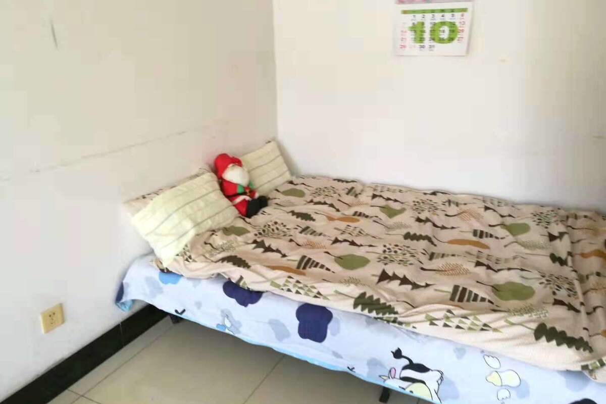 A small, single-bed room with a patterned bedspread, a plush toy, and a calendar on the wall.