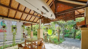Une terrasse couverte avec une table et des chaises, entourée de plantes en pot et d’un jardin.
