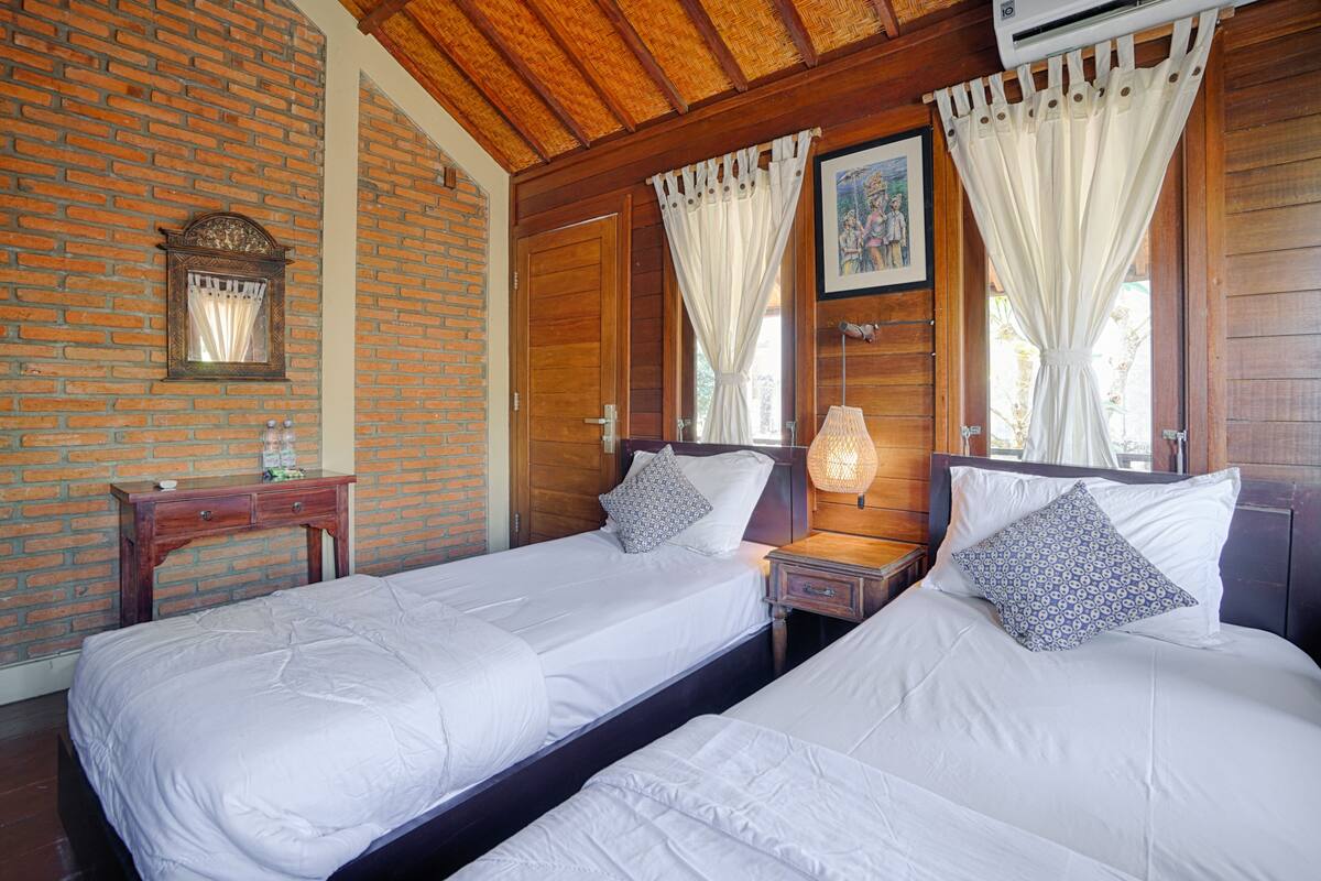 A room with two single beds, wooden walls, a brick accent wall, a wooden ceiling, and a small table with a lamp.
