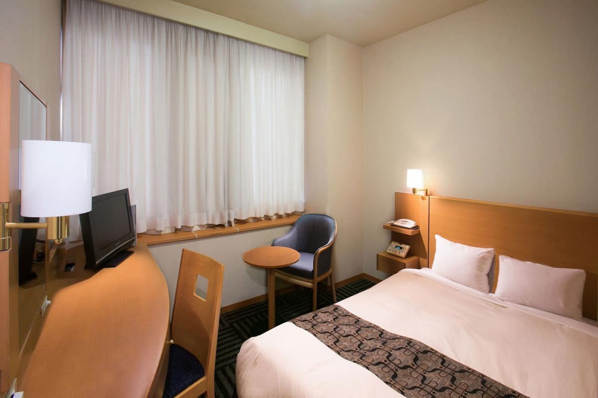 Habitación de hotel con cama, escritorio con lámpara, silla, televisor, mesita de noche con teléfono y ventana con cortinas.