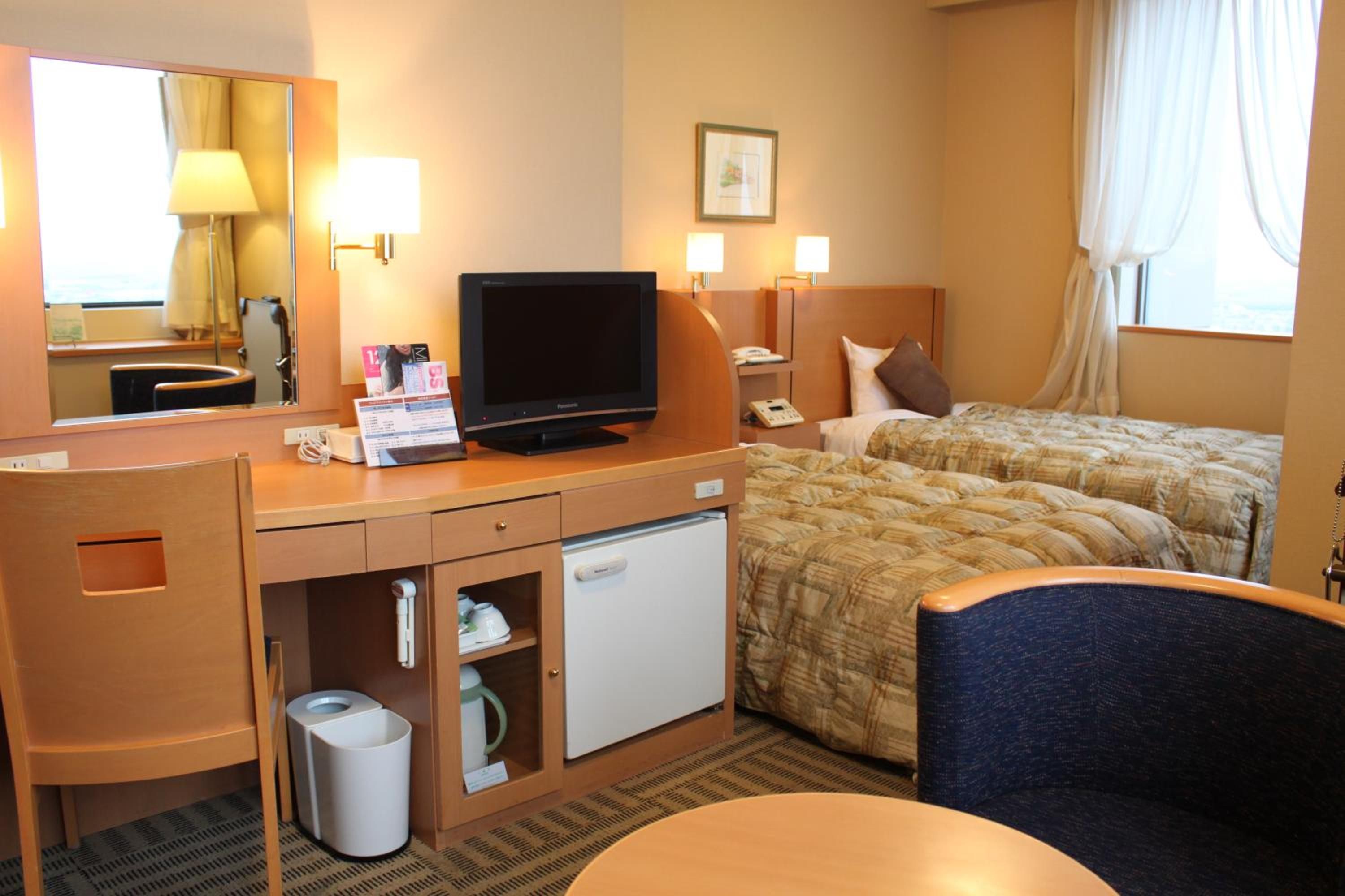 Habitación de hotel con cama, escritorio con televisor, mini refrigerador, cesto de basura y una silla.