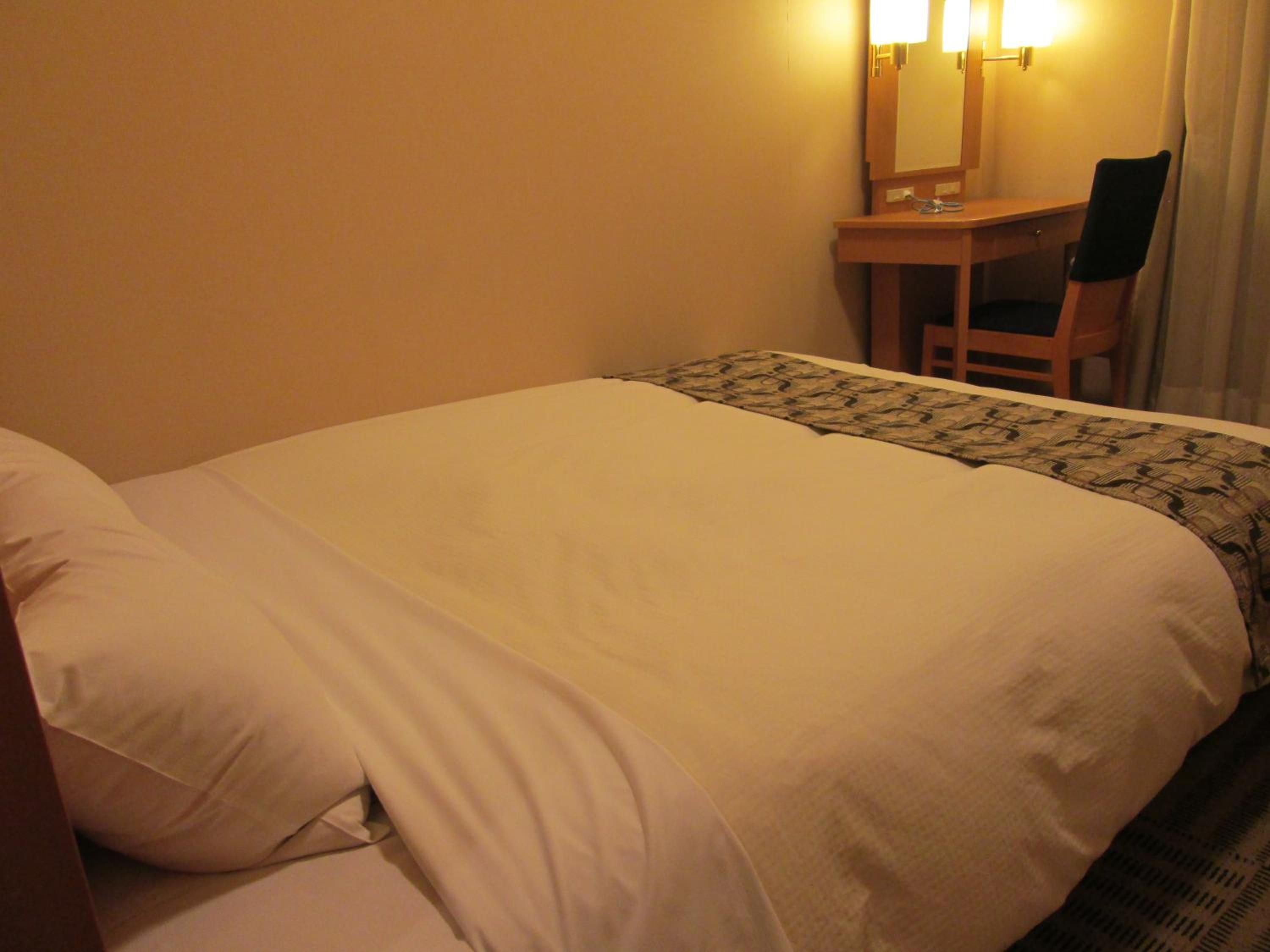 A hotel room with a bed, a desk, a chair, and a lamp.